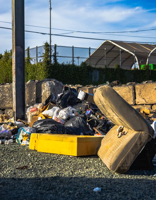 An outdoor scene showing a pile of discarded household waste, including black and yellow rubbish bags, an old, dirty car seat, and various packaging materials, all situated on a gravel surface next to a stone wall and metal fencing. In the background, there are utility poles, a hedge, and a partially visible building with a curved roof, under a blue sky with some clouds. The image appears to be taken on a bright day, with natural sunlight illuminating the waste and surrounding environment. This scene illustrates the importance of proper disposal and waste management during home removals, as managed by services such as Man with Van Norbury, who assist with household relocations, packing, and transportation logistics.