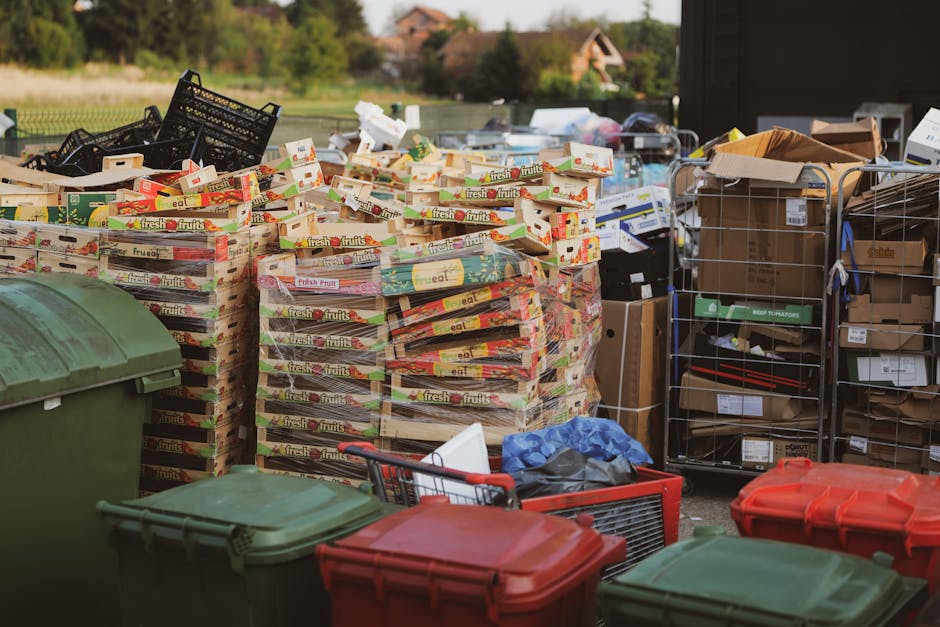 An outdoor scene depicts a large pile of emptied cardboard fruit and vegetable boxes, mainly stacked on wooden pallets, situated near a grey utility door on a property. To the right, several metal wire cages and boxes are filled with various packaging materials, including brown paper and cardboard, and some contain small appliances or household items. In the foreground, multiple coloured plastic bins with lids, such as green, red, and grey, are visible on the pavement. A shopping cart with miscellaneous small items and a blue plastic bag is placed nearby. In the background, a modern van with sliding doors open is partially visible, loaded with more boxes and packing materials, indicating a home relocation or removal process. This scene is associated with professional removals and packing within the context of furniture transport and logistics, as might be handled by Man with Van Norbury, supporting house moves and moving logistics in the Norbury area.