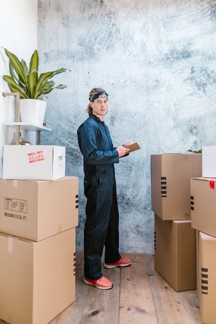 A man dressed in navy-blue work overalls and a headband stands inside a room with a textured grey wall, surrounded by several large cardboard boxes, some labeled with handling instructions. He is holding a clipboard or notepad and appears to be inspecting or taking notes during a home relocation process. The boxes are stacked on a wooden floor, with one containing a white plastic bag, and others sealed with packing tape. To the left, there is a white shelf holding a potted plant with broad green leaves. The setting suggests a packing and moving scenario, with the man likely part of a professional removals team such as Man with Van Norbury, preparing household items for transport, demonstrating a typical loading or inspection stage in furniture transport and removal services.