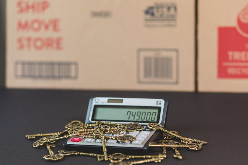A close-up view of a calculator displaying the number 749800, surrounded by a scattered pile of small brass keys with ornate bows and teeth, placed on a dark surface. In the background, there are several cardboard boxes with red and blue printed labels indicating packaging and storage, aligned in a row on a light-colored surface. The scene is indoors, with natural lighting highlighting the textures of the keys and the smooth surface of the calculator, reflecting typical steps involved in home relocation, such as packing, inventory management, and planning for furniture transport by a professional moving service like Man with Van Norbury.