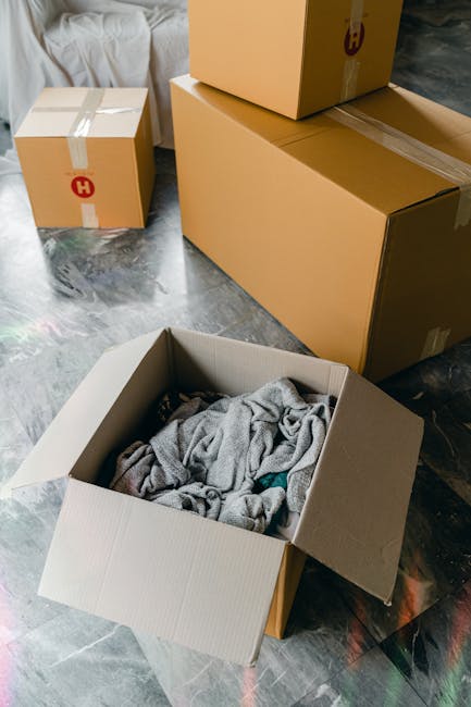 Inside a residential property, several cardboard boxes of various sizes are positioned on a gray concrete floor. One open box contains crumpled gray fabric or towels, indicating packing materials. Two larger sealed boxes, one with visible packing tape, are stacked or placed nearby, likely containing household items for home relocation. In the background, a sofa with white fabric upholstery is partially visible, suggesting a living space. The scene appears to be in the midst of a moving process, with objects prepared for transport or unpacking. The overall environment is well-lit with natural or ambient lighting, emphasizing the packing and loading activities typical of furniture transport or house removals. This image is representative of services offered by Man with Van Norbury, highlighting aspects of packing, moving logistics, and furniture transport during a residential move.