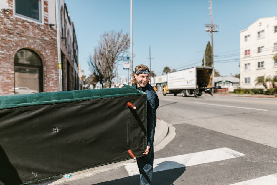 A person with light brown hair tied back, wearing a dark jacket and a patterned headband, is outside on a street in Norbury, London, during daylight. They are smiling and carrying a large, rectangular item wrapped in black shipping film or plastic, with orange handles visible on each side. The individual appears to be part of a home relocation or furniture transport process managed by Man with Van Norbury, which specializes in removals services. Behind them, there is a brick building with an arched window on the left, and on the right, a white residential building with balconies. The street features a pedestrian crossing, with a moving van or truck in the background, actively loading or unloading. Visible equipment includes the black protective wrapping on the furniture, and the person's grip on the handles suggests they are engaged in the loading or unloading process. The environment is urban, with power lines and street signs overhead, and the overall scene captures a typical furniture transport and packing stage of a house removal operation in progress.