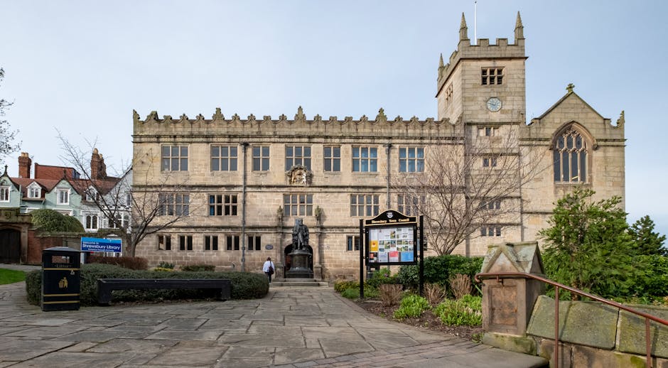 A large historic stone building with gothic architectural features, including tall pointed towers and a clock on one tower, set against a clear sky. In front of the building, there is a paved walkway with a black bench, a black waste bin, and an informational display board, all surrounded by some greenery and shrubbery. The scene appears to be part of a residential or public area, with trees and houses visible on the left side of the image. The overall setting suggests a peaceful environment suitable for community activities or local landmarks, which could be relevant to house removals or moving services in the Norbury area. The presence of the emblem and signage indicates that this is Norbury Library, a historic building, which is often associated with community and residential relocations. The image captures the exterior view, including details of the stonework, windows, and the surrounding landscape, reflecting the visual context of a typical house removal or moving logistics environment managed by Man with Van Norbury.