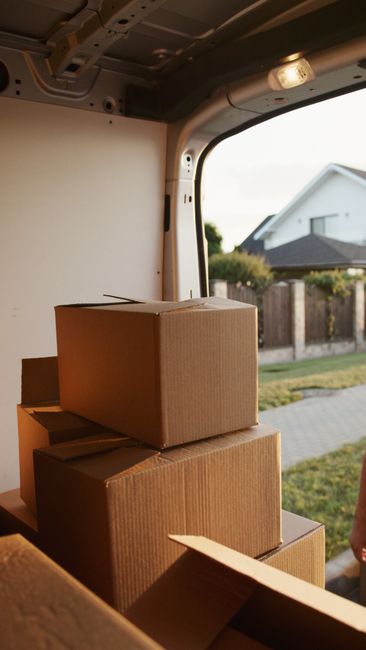 Inside the cargo area of a van used for home relocation by Man with Van Norbury, several cardboard boxes of varying sizes are stacked near the open rear door. The boxes are sealed with packing tape, and some are partially open or tilted. The van's interior roof and walls are visible, with metal and plastic components supporting the load. Outside the open door, a residential street with a paved sidewalk, a wooden fence, and a house with a pitched roof and white exterior are seen in natural daylight, indicating a daytime loading process as part of furniture transport for house removals.