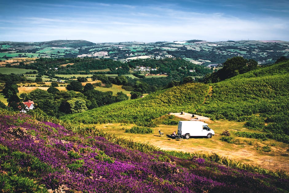 A white moving van parked on a grassy clearing amidst rolling green hills and lush agricultural fields, with rows of crops and vibrant purple heather in the foreground. The van is positioned on a dirt area and is surrounded by outdoor furniture, including lounge chairs and a small table, likely for resting during a house removal or later unpacking. The background features extensive farmland, scattered trees, and a partly cloudy sky, indicating a rural setting suitable for home relocation activities. The scene captures the outdoor loading or unloading process, with no visible personnel, but the presence of the van and surrounding materials suggests ongoing packing and furniture transport as part of a relocation service offered by Man with Van Norbury, supporting the logistics of house removals and associated moving services.