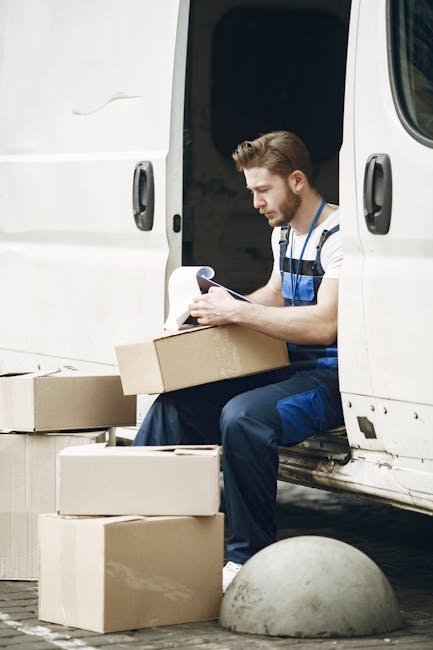 Inside the cargo area of a van used for home relocation by Man with Van Norbury, several cardboard boxes of varying sizes are stacked near the open rear door. The boxes are sealed with packing tape, and some are partially open or tilted. The van's interior roof and walls are visible, with metal and plastic components supporting the load. Outside the open door, a residential street with a paved sidewalk, a wooden fence, and a house with a pitched roof and white exterior are seen in natural daylight, indicating a daytime loading process as part of furniture transport for house removals.