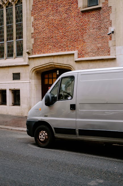 A silver commercial van parked on a street in front of a brick building with large stained glass windows and an arched doorway. The van is positioned close to the building's entrance, with the driver's side visible, showing the side mirror and door handle. The environment appears to be a residential or historic area, with the van likely used for local home relocation or furniture transport, as part of the loading process for a house move. Man with Van Norbury may be involved in this type of furniture and belongings removal, utilising the van to facilitate packing, loading, and transportation of household items. The scene captures a typical moment in the house removals process, emphasizing the importance of careful loading and transport of furniture and boxes, with the van ready for the next stage of the removal service.