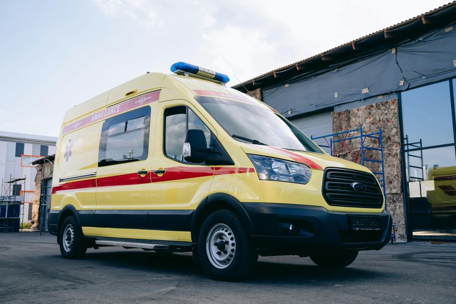 A yellow Ford ambulance van with red and purple medical markings parked outdoors on a paved surface adjacent to a building under renovation. The vehicle features a blue emergency light bar on the roof, black side mirrors, and steel wheels. The building behind has blue tarpaulin covering parts of the structure and scaffolding for ongoing work. The scene depicts a typical setting for home relocation and furniture transport involving loading or unloading supplies or equipment, with visible surroundings suggesting an area prepared for moving activities. The environment is well-lit with natural daylight, and the overall context relates to transportation and logistical support in house removals, as provided by Man with Van Norbury’s services.