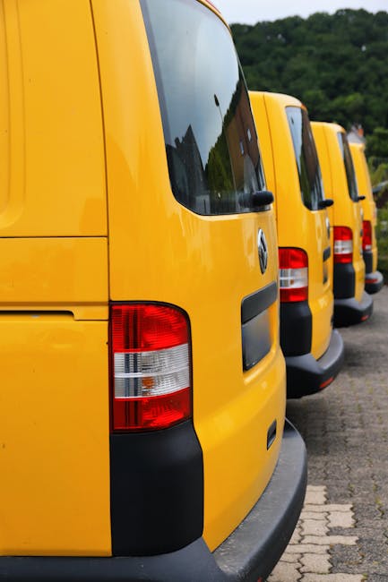 A row of three yellow vans are parked side by side on a paved surface, with the rear ends facing the camera. The vans are all Volkswagen models, featuring large rear windows and red and white tail lights. The area appears to be an outdoor loading zone near a residential or commercial building, with a slight incline and some greenery visible in the background. The environment is well-lit, suggesting daytime, and the vans are positioned close together, ready for a home relocation or furniture transport process. This parking setup reflects the types of vehicles used by Man with Van Norbury for efficient material handling during removals, packing, and moving services.