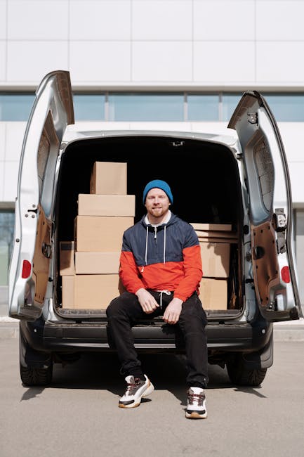 A man with a beard, dressed in a blue beanie, navy and red jacket, black pants, and white sneakers, is sitting on the open rear door of a white van parked outside a modern building with a glass facade. The van's cargo area contains several cardboard boxes of varying sizes, positioned on the floor and stacked neatly. The boxes are sealed with packing tape, and some are wrapped in plastic for protection. The loading area is well-lit by natural daylight, with the boxes clearly visible inside the vehicle. The scene depicts a home relocation or furniture transport process, with the man representing a professional removals service, such as Man with Van Norbury, preparing for a house move or packing and moving operation involving a small van suitable for household items.