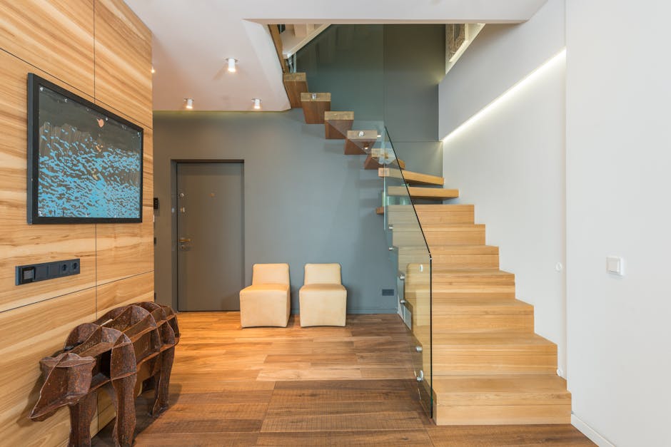Interior view of a modern residential property showing a wooden staircase with glass balustrades positioned against a white wall. The staircase extends upward to a second-floor landing, with visible steps made of light-colored wood. At the base of the stairs, on the ground floor, there are two small beige upholstered chairs placed near a blue-gray door. To the left of the image, a wall panel made of light wood features a mounted flat-screen television displaying a blue abstract pattern. Below the television, there are several electrical outlets and switches on the wall. Also visible are two decorative wooden animal sculptures, possibly deer, positioned on the wooden floor near the wall. The space is well-lit with ceiling spotlights, and the overall environment is tidy and minimalistic, suitable for home relocation or furniture transport considerations, with the setting indicating a recent or ongoing move by a professional removals service such as Man with Van Norbury.