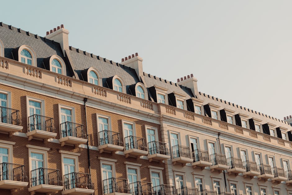 Photograph of the upper floors of a multi-storey residential building with a beige and brown facade, featuring rows of windows and small black metal balconies. The building's roof has dormer windows with rounded tops, and decorative chimneys are visible along the roofline. The image is taken from street level with natural daylight illuminating the scene. This setting is relevant to house removals and moving services, illustrating the type of property where furniture transport and packing activities, often handled by companies like Man with Van Norbury, take place during a home relocation. Inside or near a property like this, movers typically load boxes and furniture onto vans, visible in the background or nearby, using equipment such as trolleys or blankets to protect items during the loading process.